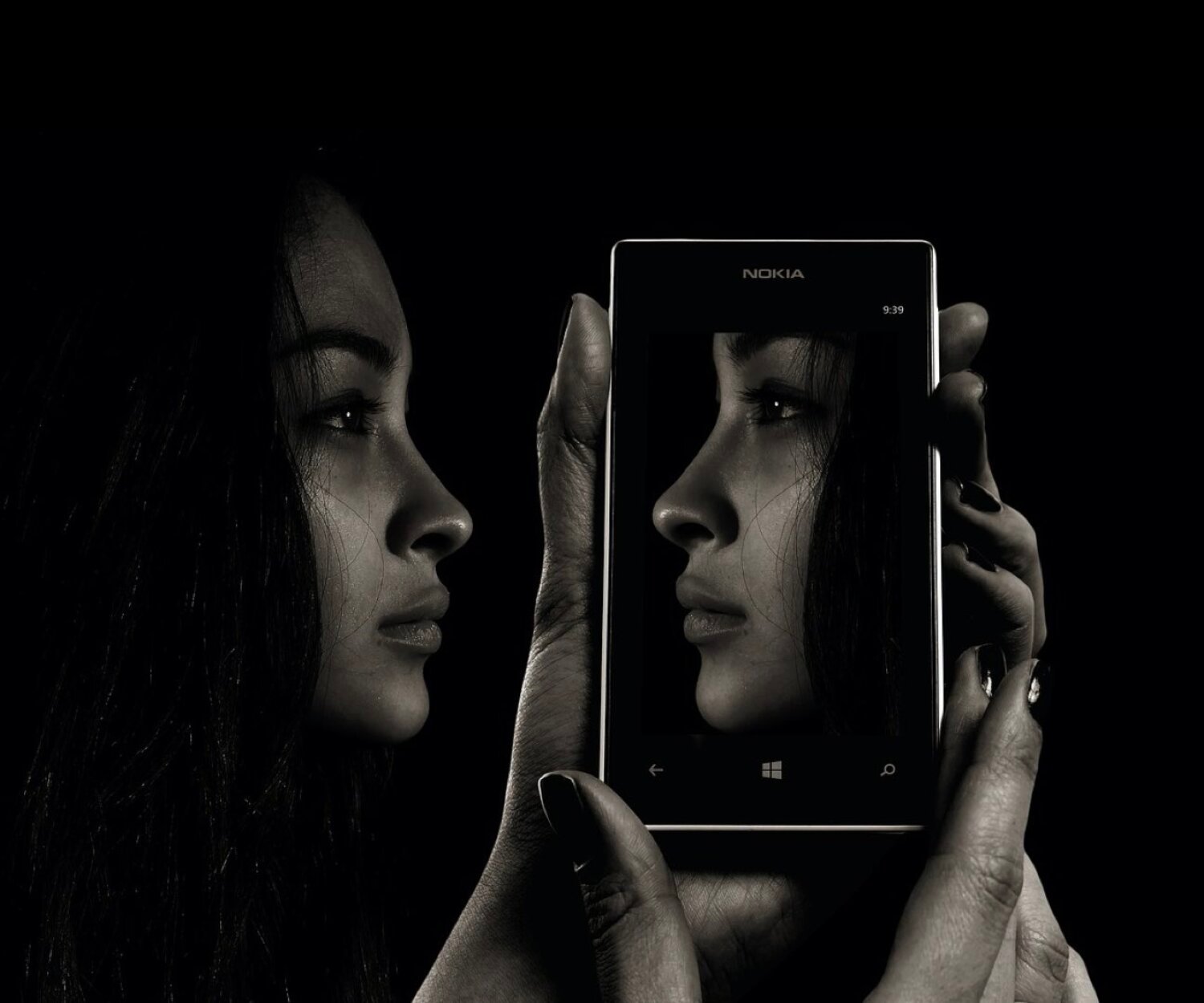 KL Tarot. A person holds a smartphone, which displays a close-up profile of their face on the screen, mirroring their actual profile visible next to it, all set against a dark background.