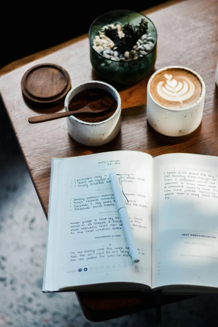 KL Tarot. Open journal with a pen rests on a wooden table alongside a latte with foam art, a bowl of cocoa powder with a wooden spoon, and a small succulent plant in a glass container.