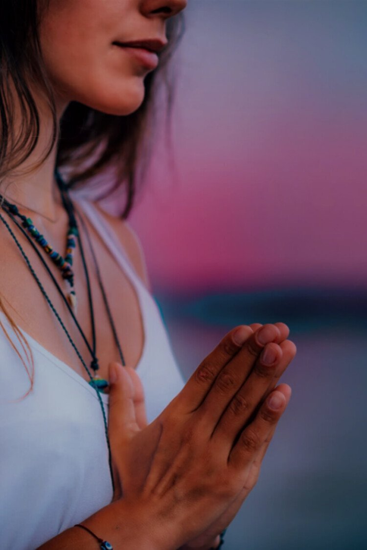 KL Tarot. A person in a white top holds their hands in a prayer position against a soft, blurred pink and blue background. Adorned with multiple necklaces, they appear deep in thought, as if contemplating the mysteries of tarot.